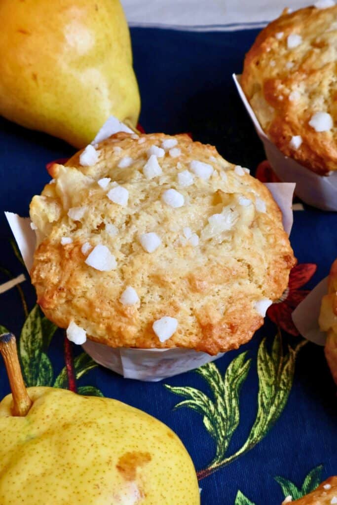 Golden pear muffin topped with coarse sugar crystals, set against a dark floral cloth and surrounded by fresh yellow pears. The muffin has a domed top and visible chunks of baked pear.