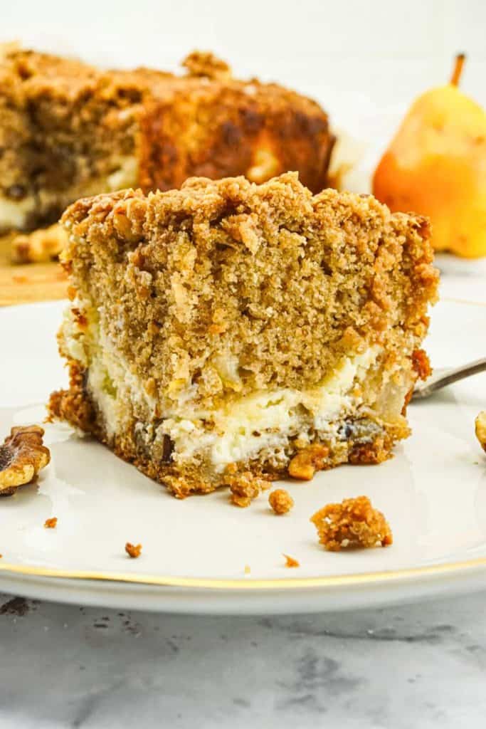 A slice of pear and walnut cream cheese coffee cake with a thick, crumbly streusel topping and creamy center, served on a white plate with scattered walnut pieces. The moist, spiced cake and visible chunks of pear make it a cozy option for fall baking ideas.