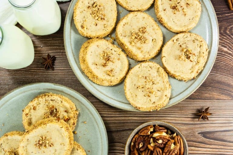 Two plates filled with round pecan shortbread cookies, each topped and edged with finely chopped pecans, set on a rustic wooden table. Nearby are glass bottles of milk, a small bowl of whole pecans, and scattered star anise for a cozy, home-baked feel.