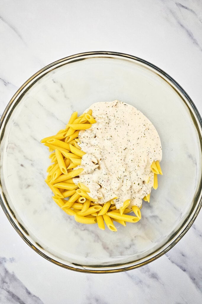 A clear glass mixing bowl containing cooked penne pasta and a generous scoop of creamy Italian hoagie-style mayo dressing, speckled with herbs and spices. The ingredients are ready to be combined on a smooth white marble surface.