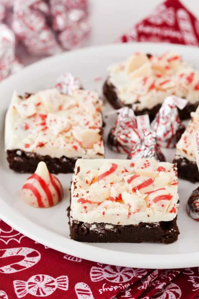 A white plate filled with rich Peppermint Bark Brownies topped with a thick layer of white chocolate and crushed peppermint candies, surrounded by red and white peppermint chocolate kisses.