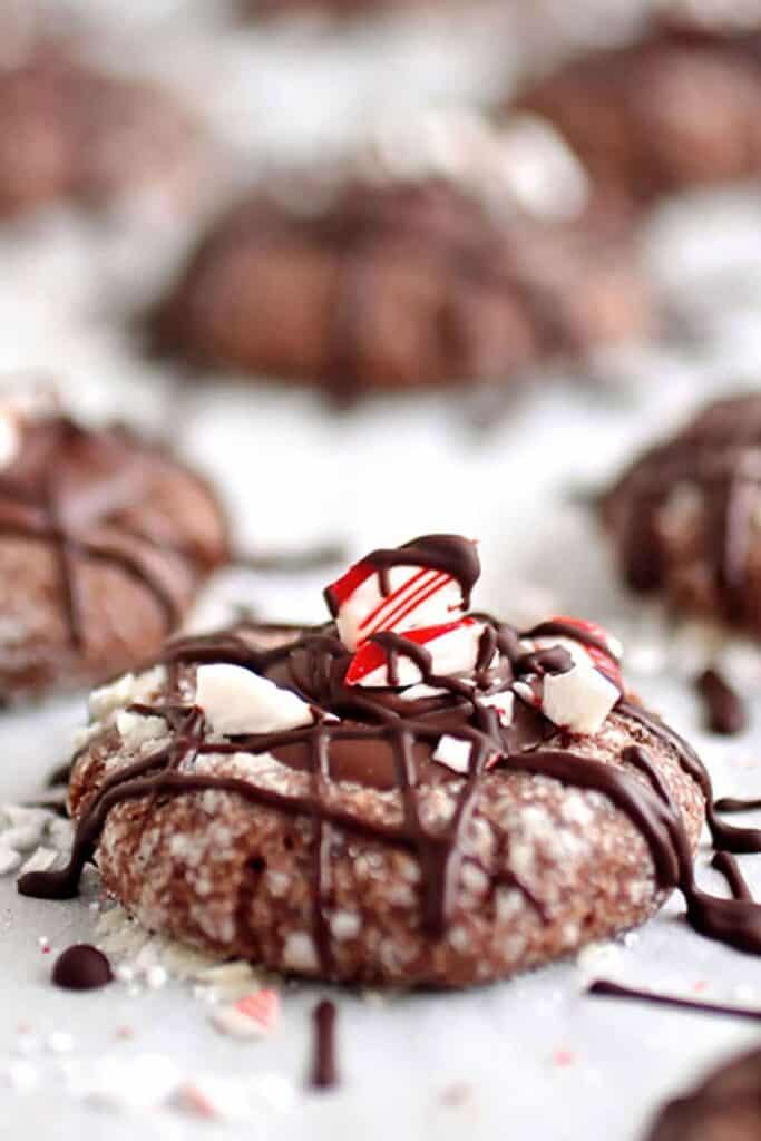 Peppermint chocolate thumbprint cookie coated in sugar, filled with melted chocolate, and topped with crushed candy canes and a chocolate drizzle. The festive cookie sits on parchment paper, surrounded by scattered peppermint pieces.