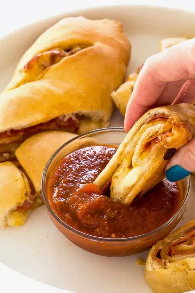 Sliced pepperoni bread with a golden, flaky crust and melted cheese inside, served on a white plate with a bowl of marinara sauce. A hand dips one warm, cheesy piece into the sauce, revealing layers of pepperoni and dough.