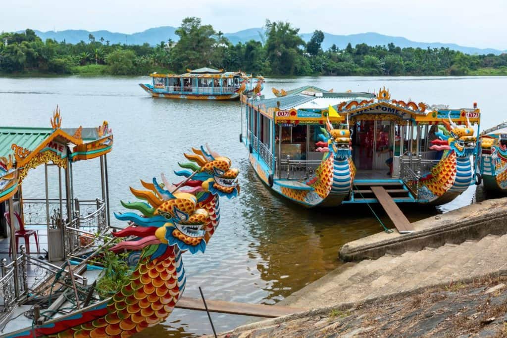 Colorful dragon boats with intricately painted dragon heads are docked along the banks of the Perfume River in Hue, Vietnam. A few boats float on the calm water, surrounded by lush greenery and distant blue mountains under a cloudy sky.