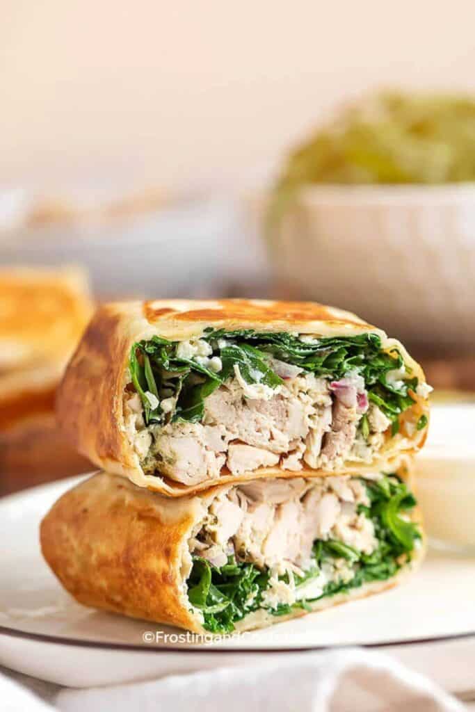 Toasted pesto chicken wrap cut in half to show a filling of tender chicken, leafy greens, red onion, and creamy cheese wrapped in a golden brown tortilla. The wrap is stacked on a white plate, highlighting an easy, protein packed meal that comes together quickly for lunch or dinner.