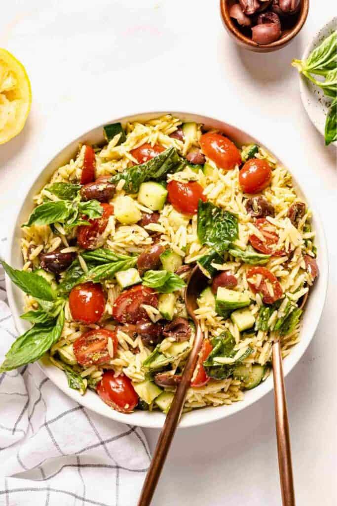 Bright and colorful pesto orzo salad served in a white bowl, featuring orzo pasta tossed with cherry tomatoes, cucumber chunks, Kalamata olives, and fresh basil leaves. Two bronze serving utensils are placed in the bowl, with lemon, olives, and basil arranged around it for a fresh, Mediterranean vibe.