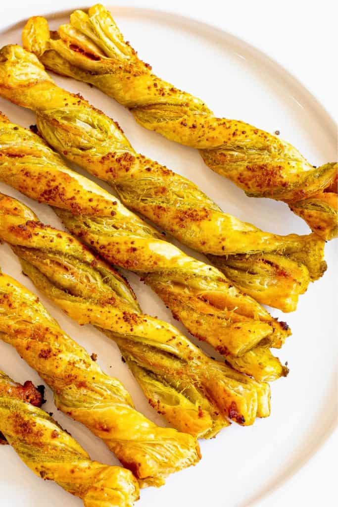 Golden brown Pesto Parmesan Cheese Straws twisted with layers of flaky puff pastry, vibrant green pesto, and melted parmesan are served on a white plate.