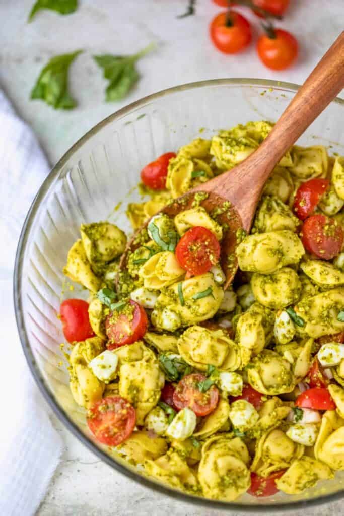 Bowl of pesto tortellini pasta salad with cheese tortellini cherry tomatoes fresh basil and mozzarella pearls tossed in green pesto sauce. Wooden spoon serving the salad in a glass bowl suggests an easy camping salad and make ahead side dish.