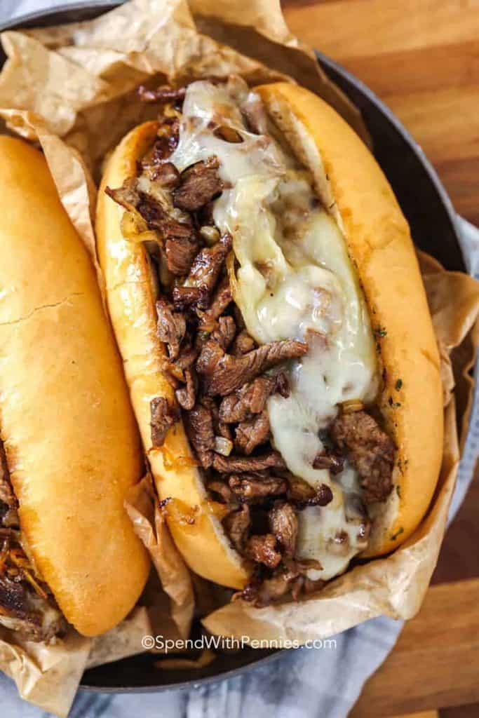 Close-up of a cheesesteak sandwich filled with sliced, seasoned steak, sautéed onions, and melted provolone cheese inside a toasted hoagie roll. Served in parchment paper, this hearty option is a classic choice for game day finger foods.