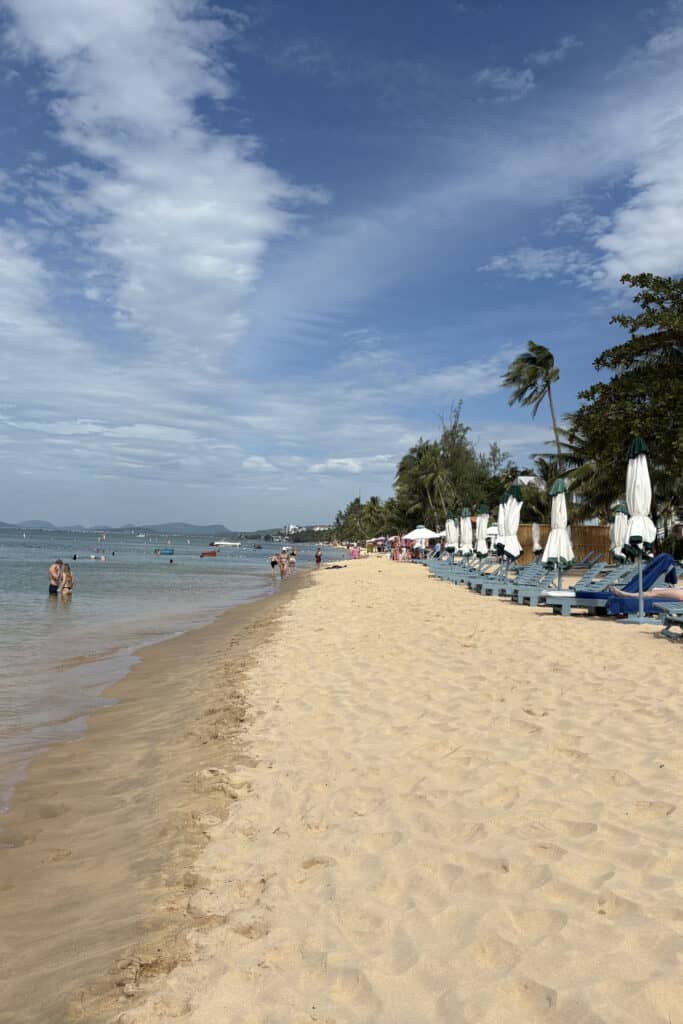 A sunny tropical Phu Quoc beach with soft golden sand, calm blue water, and rows of empty lounge chairs with closed umbrellas lining the shore. A few people wade in the shallow waves under a partly cloudy sky, with palm trees and resort-style buildings in the background.