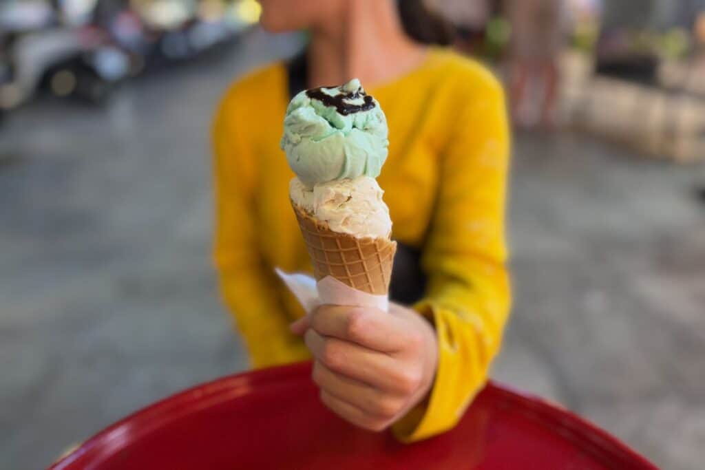 A young girl in a yellow top holds a waffle cone with two scoops of gelato—mint with chocolate drizzle on top and a creamy vanilla flavor below—while sitting at a red table on a street in Phú Quốc.