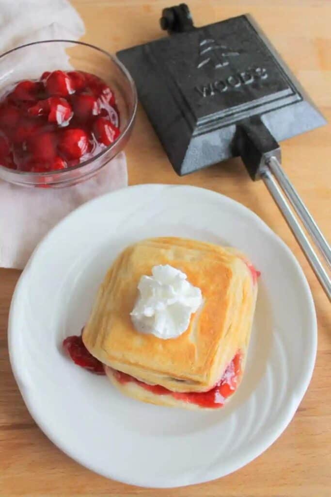 Plate with camp cooker cherry pie, topped with whip cream, beside a small bowl of cherry filling, and cast iron pie iron.