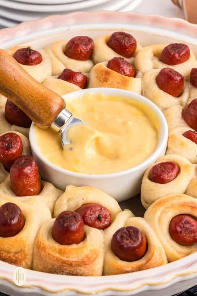 A dish of golden-brown pigs in a blanket arranged in a ring around a bowl of creamy cheese dip, with a wooden-handled spoon ready for serving. An easy finger food for Super Bowl parties that's warm, cheesy, and crowd-pleasing.