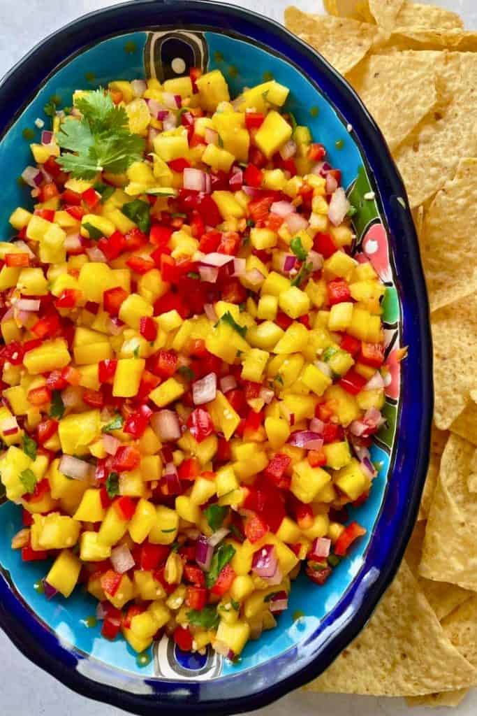 Colorful pineapple mango habanero salsa served in a vibrant blue dish, featuring diced yellow mango, pineapple, red bell pepper, red onion, cilantro, and habanero peppers. Fresh tortilla chips are placed on the side, making it a bold and fruity cold dip perfect for tailgate parties.