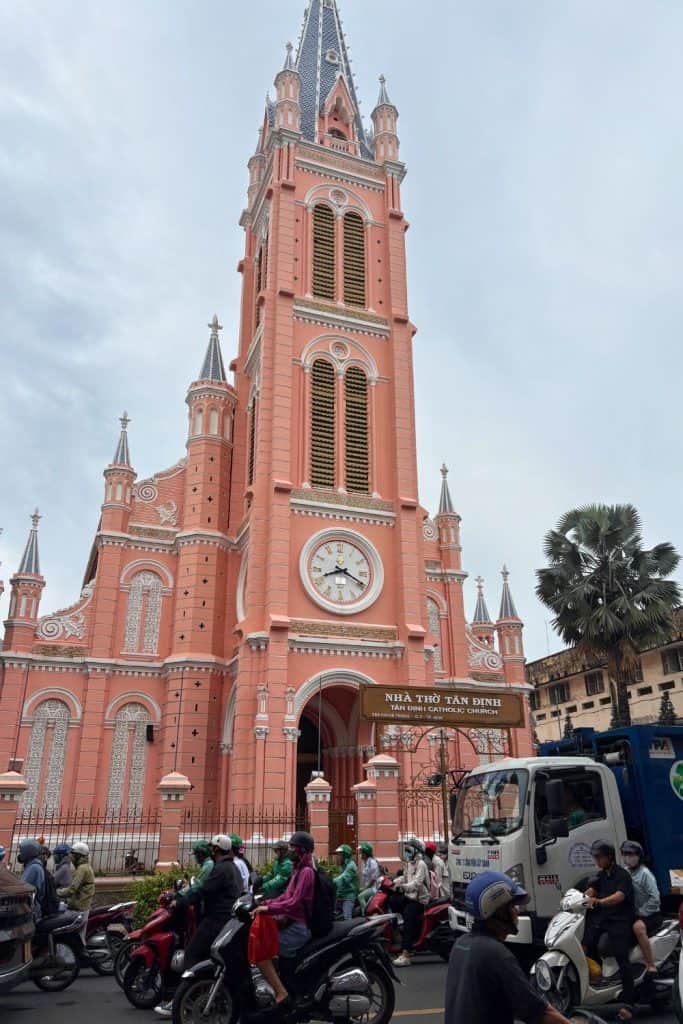The bright pink facade of Tan Dinh Catholic Church stands tall with its ornate white trim, clock tower, and pointed spires against a cloudy sky. In front of the church, a busy street scene unfolds with motorbikes, scooters, and a truck weaving through the traffic in Ho Chi Minh City.