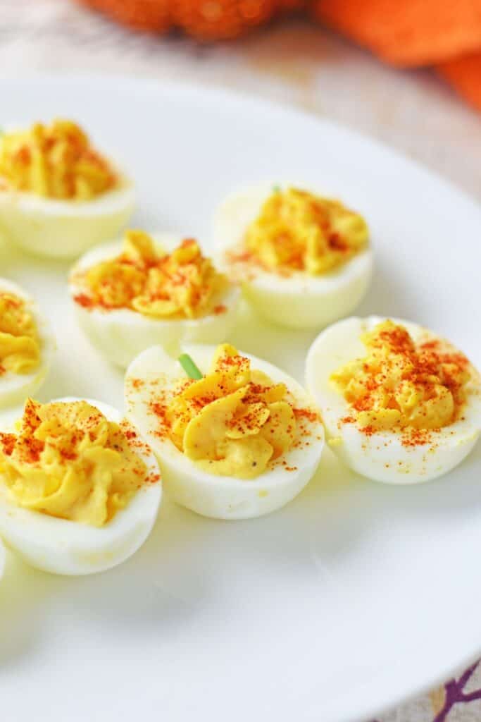 Deviled eggs in rows with creamy yellow filling dusted with smoked paprika and topped with small green chives forstems. The filling is piped into the egg whites to resemble miniature pumpkins for a festive Fall and holiday snack.