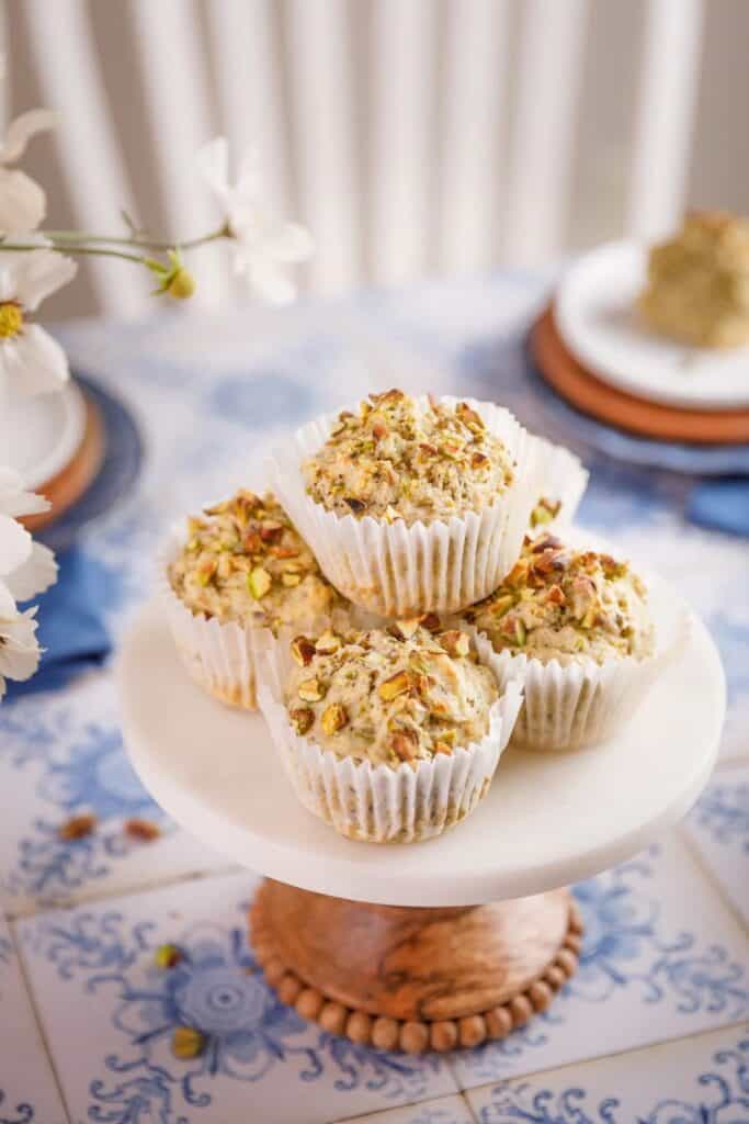 Pistachio muffins stacked on a white marble cake stand, each topped with chopped pistachios and wrapped in white paper liners. The setting features a soft, floral aesthetic with blue-and-white tile patterns and delicate tableware in the background.