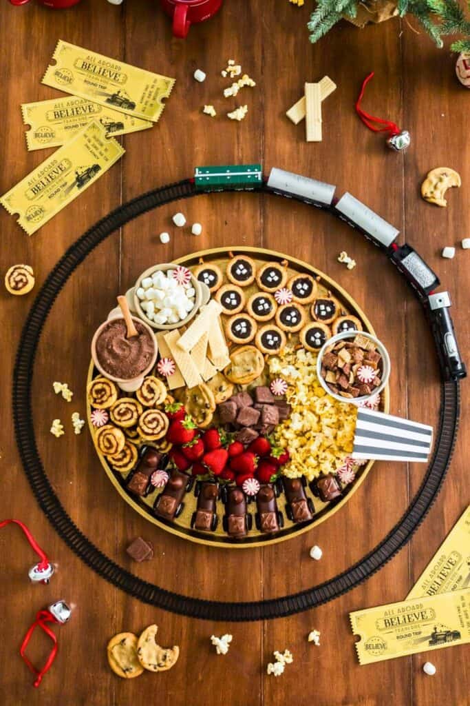 A festive snack board filled with Christmas movie night snacks, including chocolate-covered marshmallow trains, strawberries, chocolate fudge, popcorn, mini cookies, Chex mix, hot cocoa with marshmallows, and holiday candies, surrounded by a toy train track and golden Polar Express-style tickets.