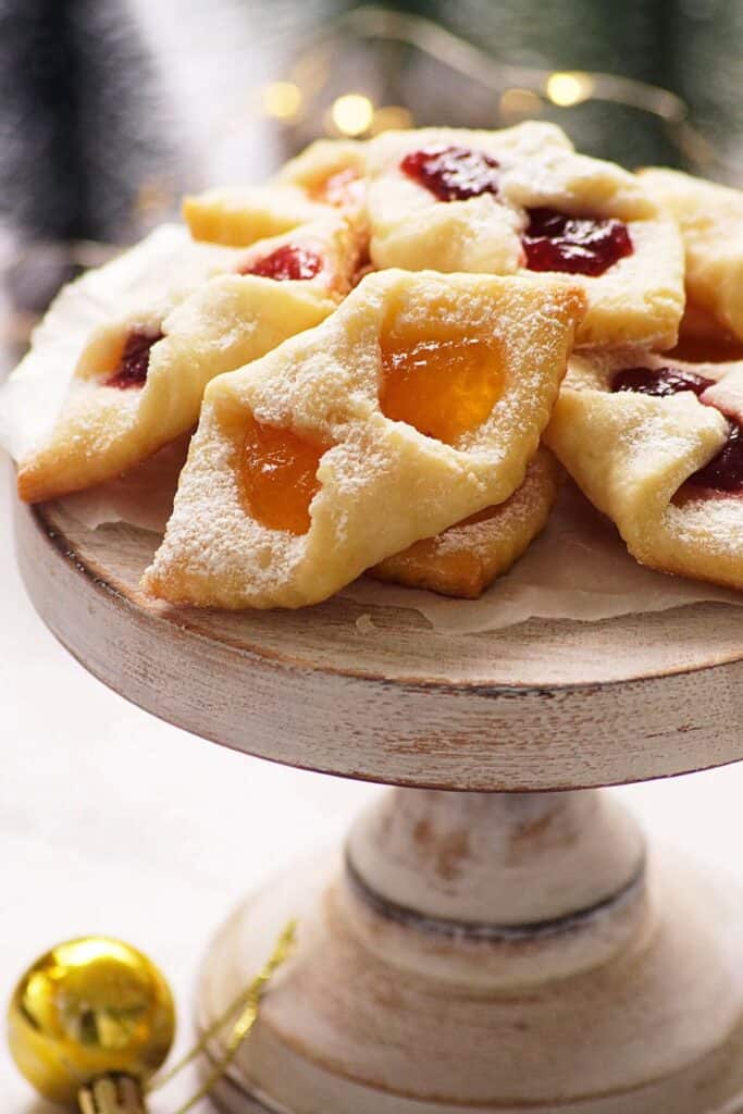 Kolaczki cookies filled with golden apricot and deep red raspberry jam are folded into diamond shapes and dusted with powdered sugar. Displayed on a rustic cake stand, these delicate Eastern European treats are a festive favorite for Christmas celebrations and cookie exchanges.