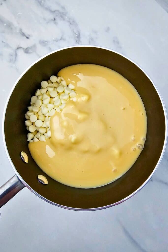 Overhead view of a saucepan filled with white chocolate chips and sweetened condensed milk, ready to be melted together for a fudge base. 