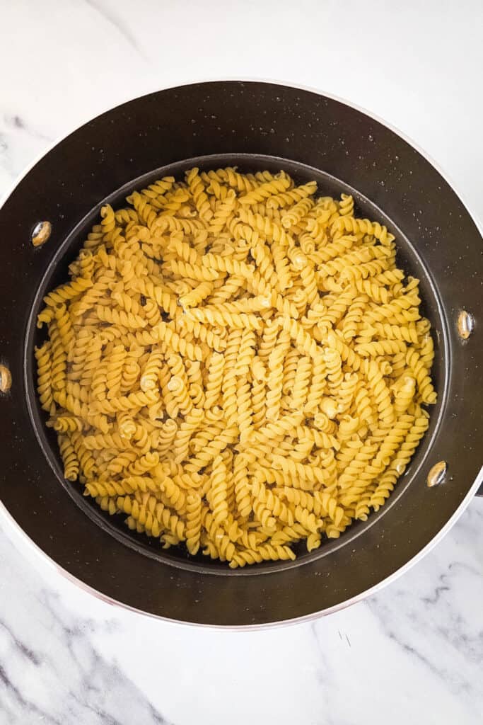 Overhead view of a pot filled with uncooked rotini pasta submerged in water, resting on a white marble countertop, ready to be boiled.