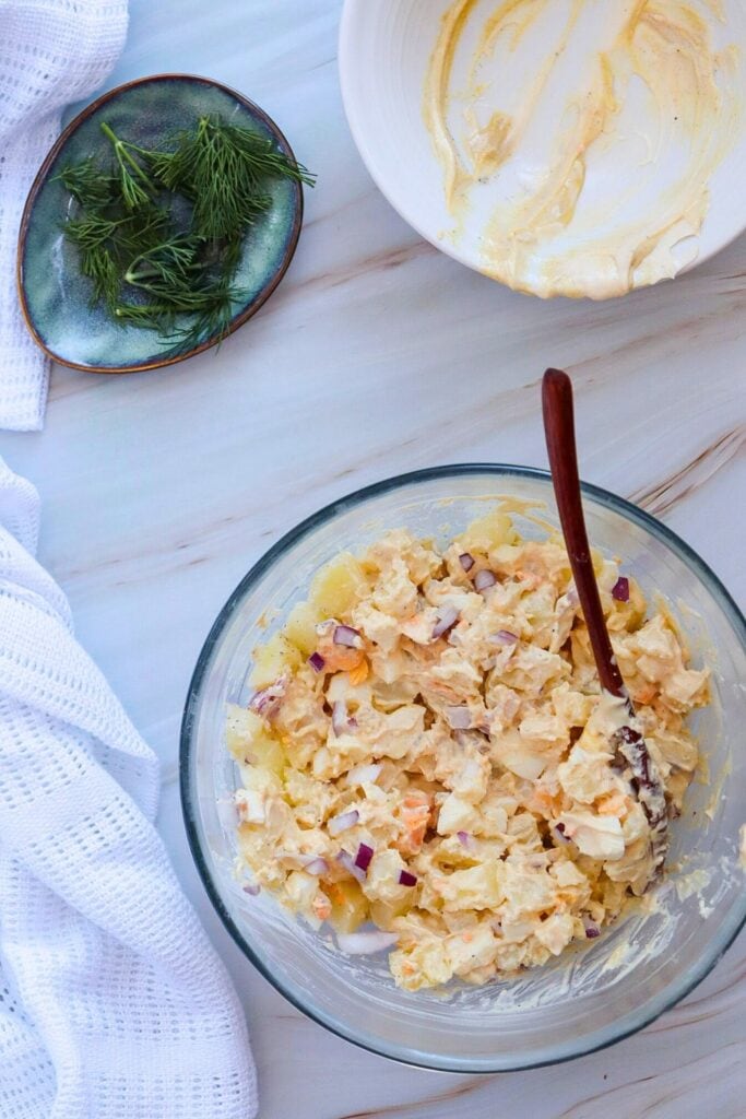 Mixing potato salad with egg and mayo dressing in a bowl, with a small oval dish of fresh dill beside it. 