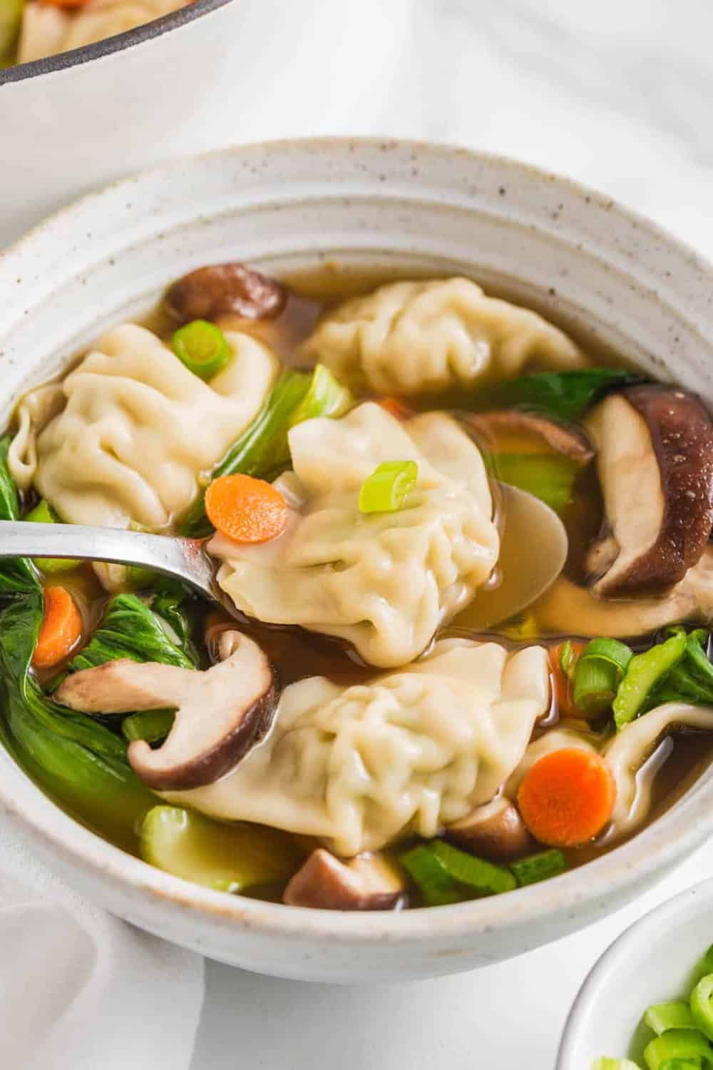 Potsticker soup with plump dumplings shiitake mushrooms carrots and leafy greens is served in a savory broth. A spoon lifts one potsticker from the colorful vegetable soup.