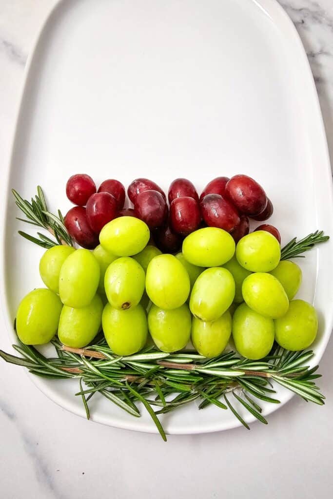 A holiday fruit and cheese platter in progress, with layers of green and red grapes arranged in a Christmas tree shape on a white oval serving tray, accented with fresh rosemary sprigs.