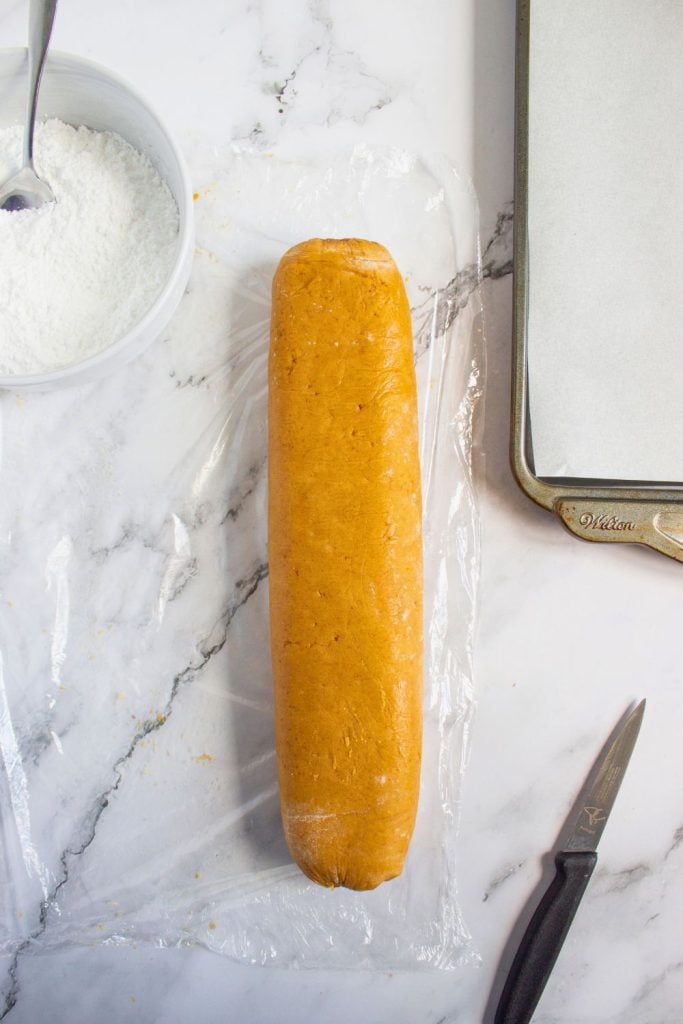 Chilled pumpkin cookie dough log unwrapped from plastic, placed on a marble surface next to a bowl of powdered sugar, a paring knife, and a parchment-lined baking sheet, ready to be sliced into cookies.