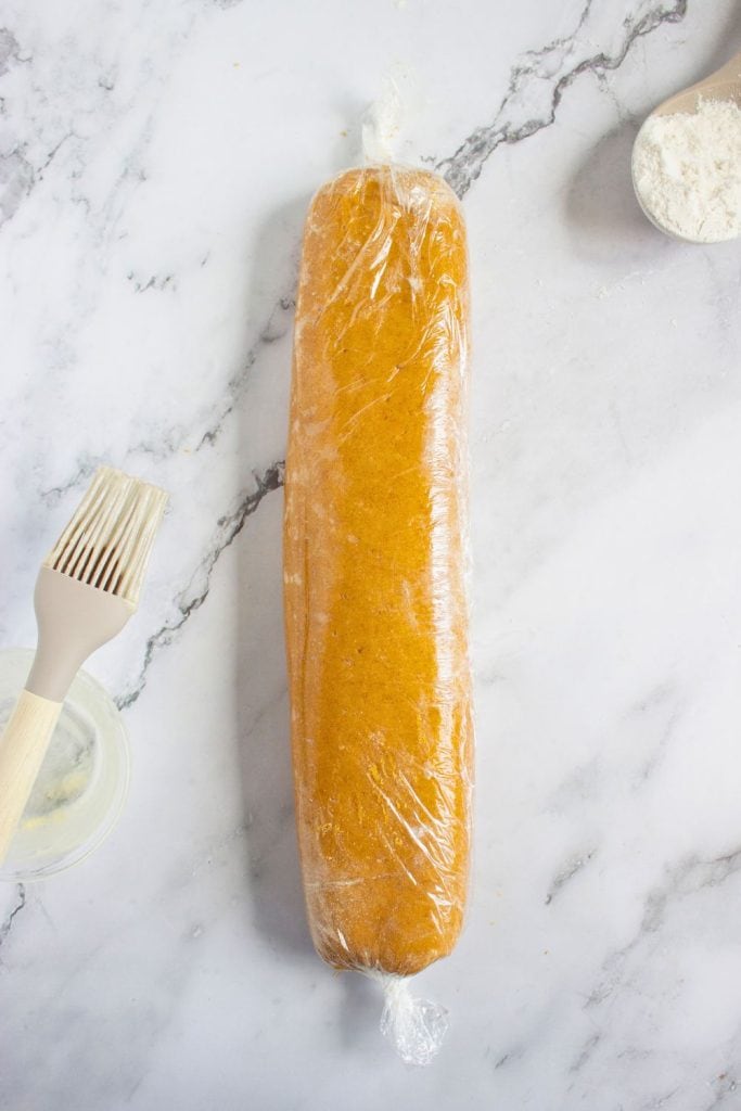 Pumpkin cinnamon cookie dough rolled into a log and wrapped tightly in plastic wrap, ready to chill or freeze. The dough rests on a marble surface next to a pastry brush, flour, and a small bowl.
