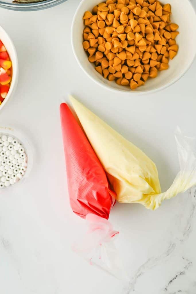 Close up of ingredients for Oreo turkeys, featuring piping bags filled with red and vanilla icing, a bowl of butterscotch chips, candy eyes, and candy corn. These colorful elements are arranged on a white surface, ready for decorating Thanksgiving-themed treats.