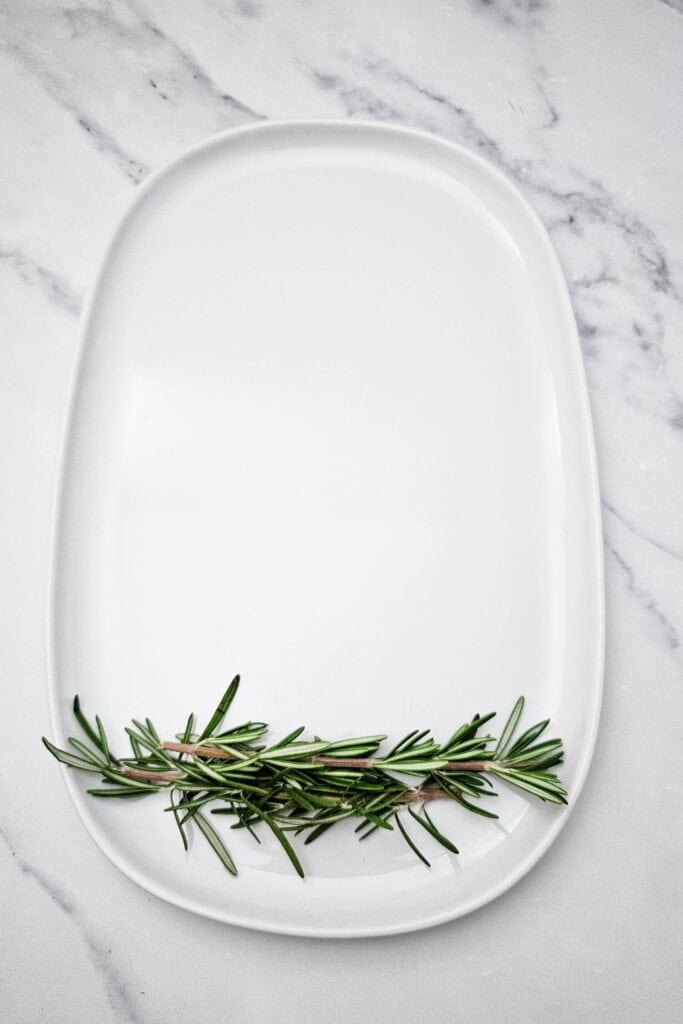 Fresh rosemary sprigs arranged along the bottom of a white oval fruit tray, creating the base for a Christmas tree shaped fruit and cheese platter.