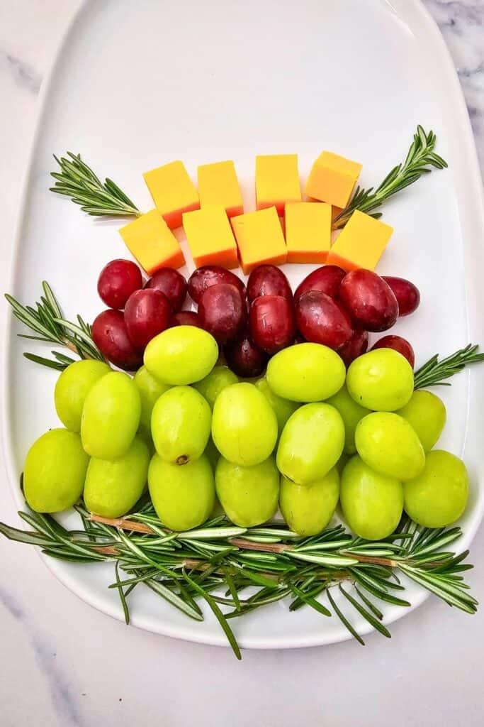 A festive fruit platter taking shape on a white oval tray, with layers of green and red grapes, bright cheddar cheese cubes, and fresh rosemary sprigs arranged to resemble Christmas tree branches.