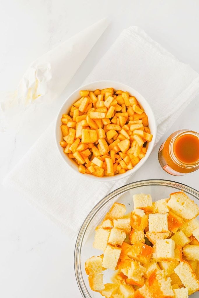 Ingredients for fall apple trifles, including a bowl of diced spiced apples, cubed pound cake in a glass bowl, a piping bag of whipped topping, and a jar of caramel sauce. The setup is arranged on a white towel over a light surface, ready for dessert assembly.