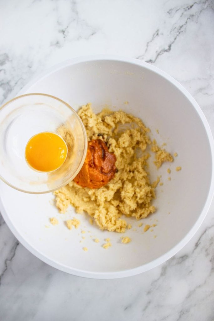Egg yolk in a glass dish being added to a mixing bowl with creamed butter, sugar, and a scoop of pumpkin purée. The ingredients are partway through preparation on a marble countertop.
