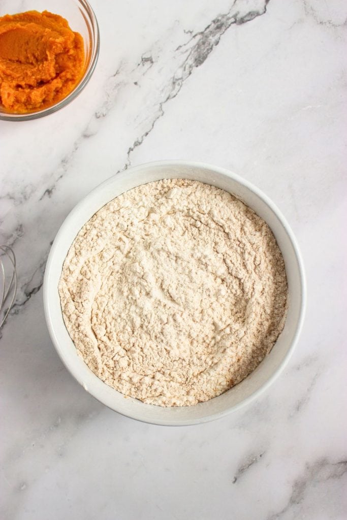 Bowl of flour mixed with pumpkin spices like cinnamon, ginger, and nutmeg, forming a light brown dry mixture. A whisk and bowl of pumpkin purée sit nearby on a marble surface, ready for the next baking step.