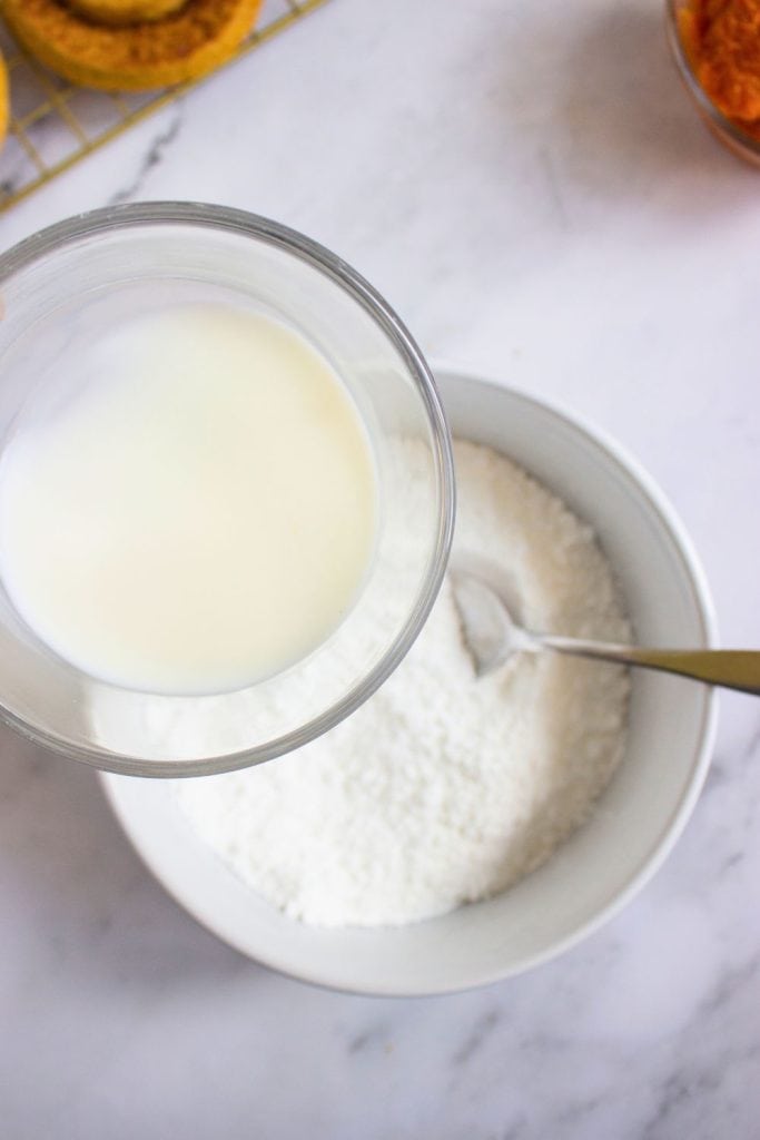 Close-up of milk being poured into a bowl of powdered sugar to make a glaze, with a spoon in the bowl and cookies visible on a cooling rack in the background. The setup rests on a marble surface, surrounded by fall baking ingredients.