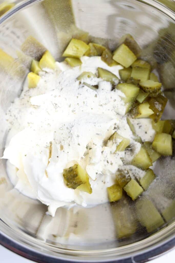 Unmixed ingredients for dill pickle dip in a stainless steel bowl, including chopped pickles, cream cheese, sour cream, and powdered ranch seasoning.