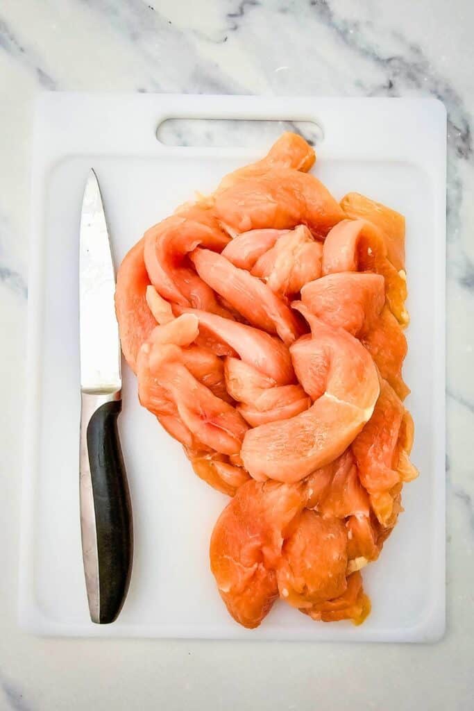 Raw sliced chicken breasts arranged on a white cutting board with a kitchen knife placed alongside.
