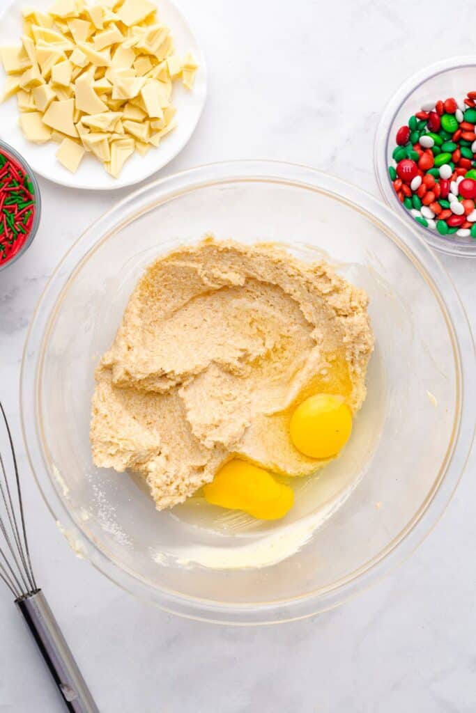 Overhead view of creamed butter being mixed in a glass bowl with two cracked eggs on top, surrounded by bowls of chopped white chocolate, red and green sprinkles, and Christmas M&Ms, ready for holiday baking.