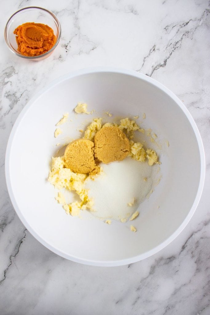 Mixing bowl with softened butter, brown sugar, and granulated sugar before creaming together. A small bowl of pumpkin purée sits nearby on a marble surface, prepping for the next baking step.