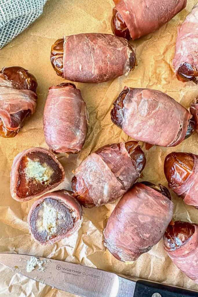 Prosciutto wrapped dates stuffed with creamy goat cheese are arranged on parchment paper after baking. Several dates are cut open to show the soft goat cheese filling inside the caramelized dates and crisp prosciutto.