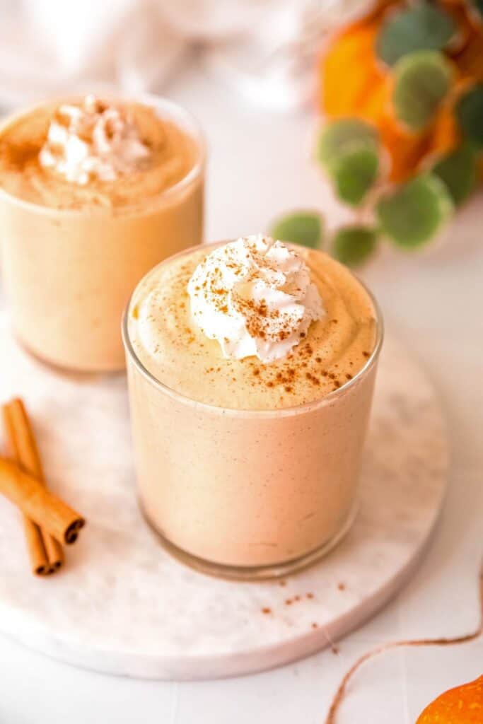 Creamy protein pumpkin fluff served in two glasses and topped with whipped cream and a dusting of cinnamon. Cinnamon sticks and a small pumpkin add a cozy fall touch around the high protein fall dessert.