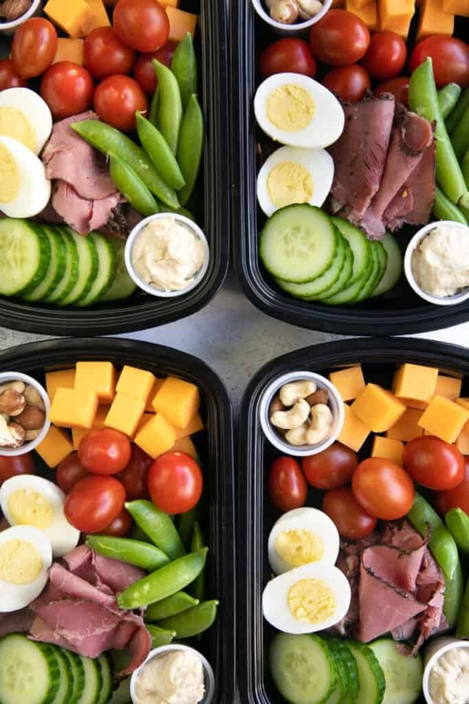 Four mini containers of protein snacks including cheese cubes, cherry tomatoes, sliced beef, boiled eggs, cucumbers, peas and dip. 