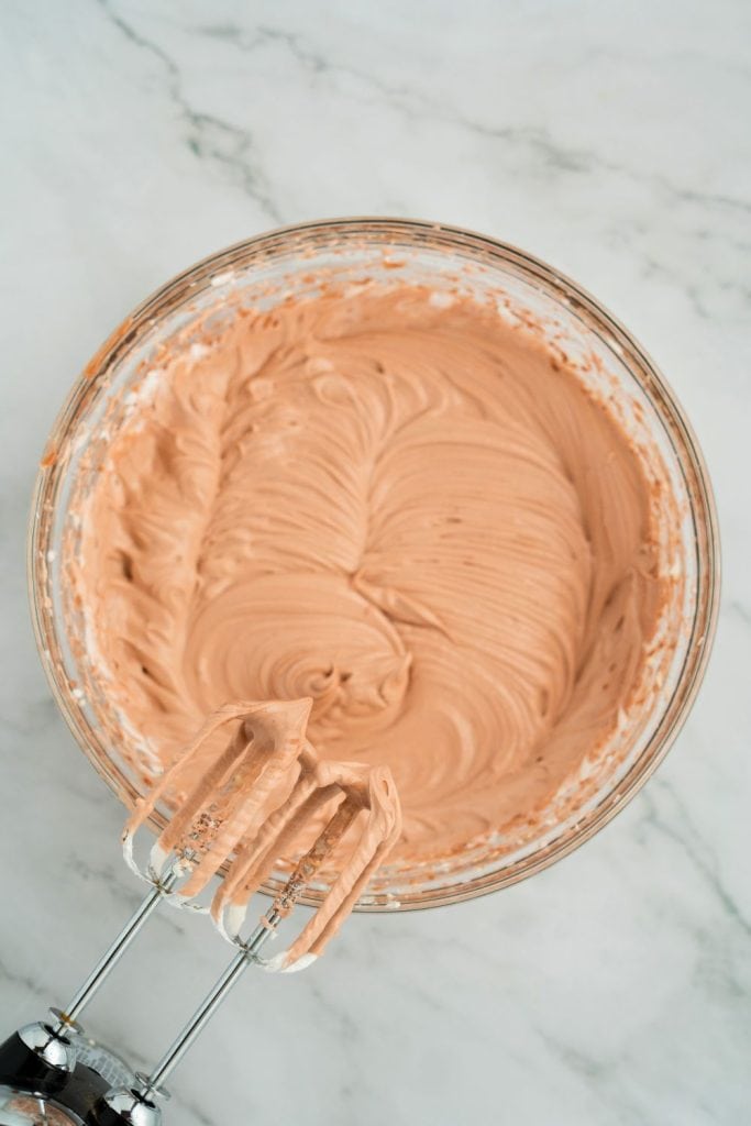 Glass bowl filled with smooth, light chocolate pudding cream cheese and cool whip mixture after being fully blended, with electric mixer beaters resting nearby on a marble countertop.