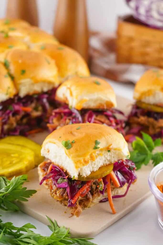 Pulled pork sliders topped with colorful red cabbage slaw, sliced pickles, and served on soft golden buns, garnished with fresh herbs and surrounded by extra pickles and parsley on a serving tray.