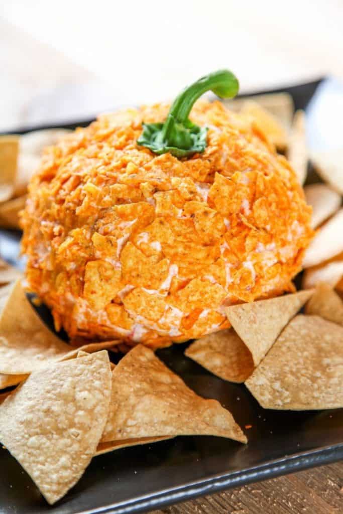 Pumpkin-shaped cheese ball coated in crushed orange chips, topped with a green bell pepper stem to mimic a real pumpkin. Surrounded by tortilla chips on a black serving tray, this festive appetizer is styled perfectly for fall gatherings or Halloween parties.