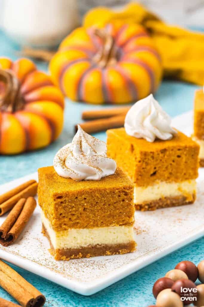 Two squares of pumpkin cheesecake bars on a plate with a dollop of whip cream on top.