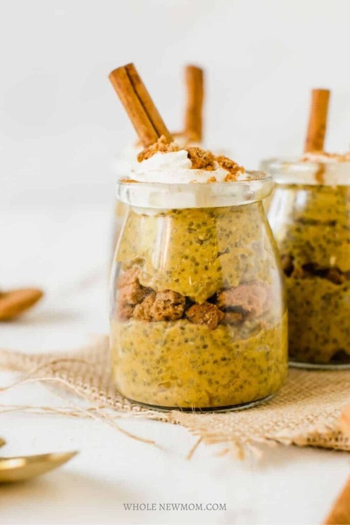 Pumpkin chia pudding layered with crumbled cookies in a small glass jar and topped with whipped cream and a cinnamon stick. The creamy orange pudding is speckled with chia seeds.