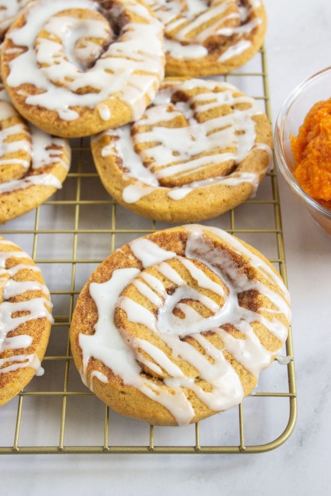 Pumpkin cinnamon roll cookies topped with white icing, cooling on a gold wire rack. A glass bowl of vibrant orange pumpkin purée sits nearby.
